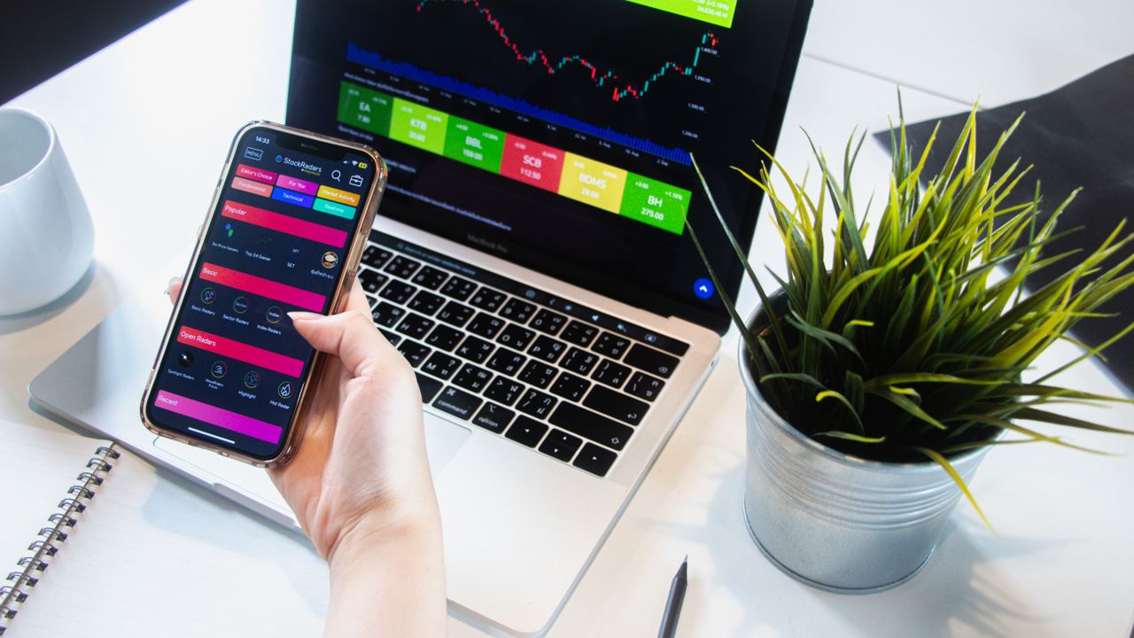 services-03 A person using a smartphone app to trade stocks with a laptop displaying market data.